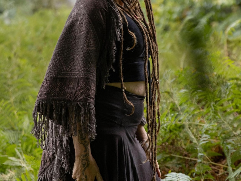 May include: A dark brown shawl with a geometric pattern and fringe detail. The shawl is draped over a black top and skirt. The image is set against a backdrop of green foliage.