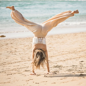 May include: A person doing a handstand on a sandy beach wearing beige pants and a white top with a lace-up back.