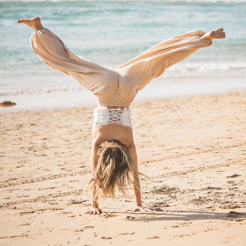 May include: A person doing a handstand on a sandy beach wearing beige pants and a white top with a lace-up back.