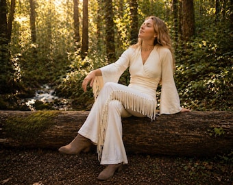 May include: Cream-colored, long-sleeved top with flared sleeves and matching pants with fringe detail. The outfit is worn with brown boots. The person is sitting on a log in a forest setting with sunlight filtering through the trees.