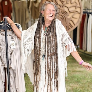 Op de afbeelding: Een vrouw met lange dreadlocks glimlacht terwijl ze een witte gehaakte sjaal met franjes vasthoudt. Ze staat voor een kledingrek met verschillende kledingstukken die eraan hangen.