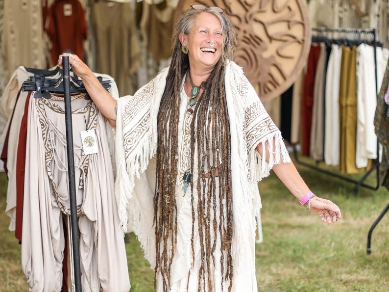Op de afbeelding: Een vrouw met lange dreadlocks glimlacht terwijl ze een witte gehaakte sjaal met franjes vasthoudt. Ze staat voor een kledingrek met verschillende kledingstukken die eraan hangen.