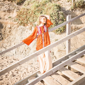 May include: A woman wearing a long-sleeved orange and purple patterned dress with a white skirt and yellow sunglasses, standing on a wooden staircase.