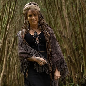 May include: A woman in a black top with a lace-up front, a brown fringed shawl, and a head wrap. She has long dreadlocks and is in a forest setting.