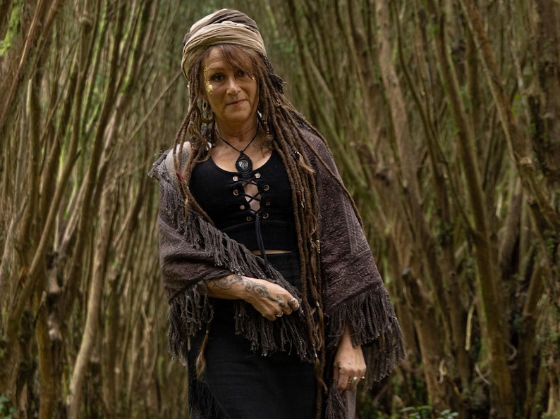 May include: A woman in a black top with a lace-up front, a brown fringed shawl, and a head wrap. She has long dreadlocks and is in a forest setting.