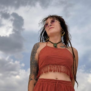 May include: A rust-coloured crop top featuring a high neckline and a fringe hemline. The garment is sleeveless with thin straps and is paired with matching rust-coloured bottoms that have an elastic waistband and a drawstring tie at the centre.