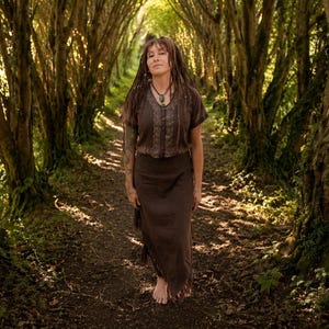 May include: A woman in a brown top and skirt stands on a path in a forest. The clothing has a textured design. The path is surrounded by trees with sunlight filtering through the leaves, creating a natural, earthy scene.