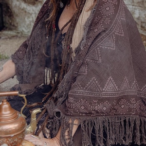 May include: A brown shawl with a geometric pattern and fringe. The shawl is draped over the shoulders. The shawl's design includes diamond and triangle shapes. The color is a deep brown.