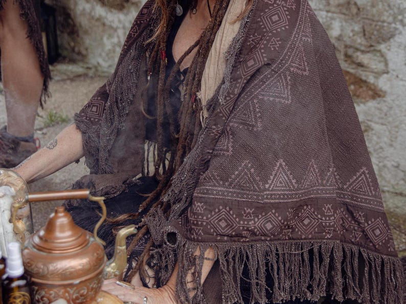 May include: A brown shawl with a geometric pattern and fringe. The shawl is draped over the shoulders. The shawl's design includes diamond and triangle shapes. The color is a deep brown.