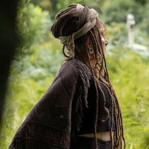 May include: A person wearing a dark brown shawl with a geometric pattern and fringe. They have long dreadlocks and a head wrap. The background is a blurred green landscape.