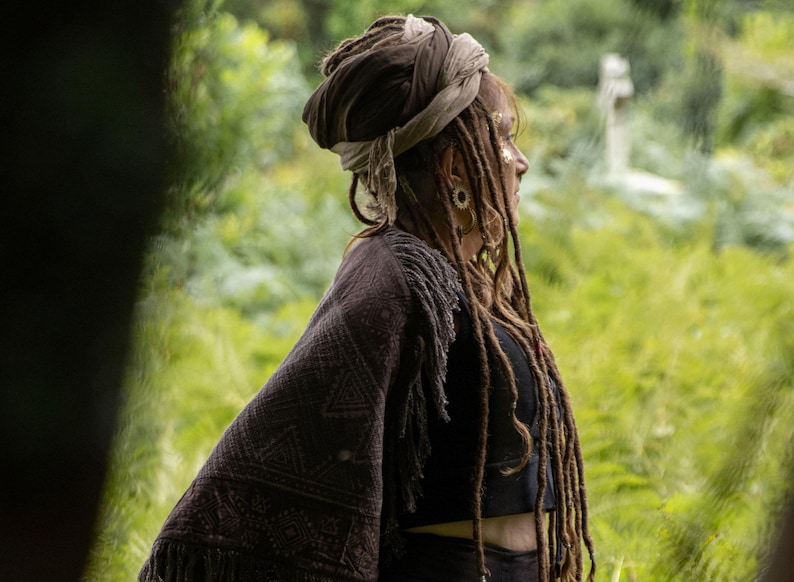 May include: A person wearing a dark brown shawl with a geometric pattern and fringe. They have long dreadlocks and a head wrap. The background is a blurred green landscape.