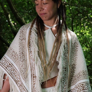 Op de afbeelding: Een witte katoenen shawl met bruine blokgeprinte ontwerpen en franjes. De shawl wordt gedragen door een persoon met lange bruine dreadlocks.