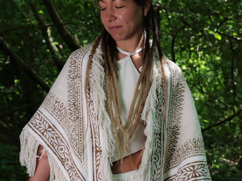Op de afbeelding: Een witte katoenen shawl met bruine blokgeprinte ontwerpen en franjes. De shawl wordt gedragen door een persoon met lange bruine dreadlocks.