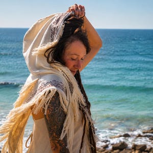 May include: Cream-colored hooded shawl with fringe and patterned trim. The shawl is worn outdoors with the ocean in the background. The trim features a geometric design in brown and red. The shawl is part of a bohemian clothing collection.