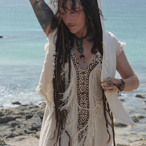 Op de afbeelding: Een crèmekleurige jurk met franjes en een geometrisch bruin patroon aan de voorkant. De jurk heeft een capuchon en wordt gedragen aan zee. De persoon draagt een ketting met groene stenen en meerdere ringen.