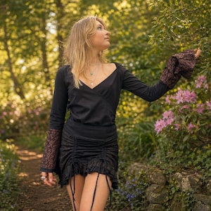 Puede incluir: Conjunto negro de top de manga larga y falda a juego. El top tiene cuello en V y mangas con puños de encaje marrón. La falda es fruncida con detalles de cordón. El conjunto se usa al aire libre.