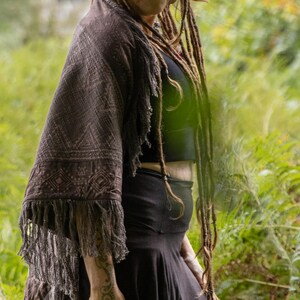 May include: A dark brown shawl with a geometric pattern and fringe is worn over a black top and skirt. The image is set in a green, leafy environment. The shawl's pattern is visible.