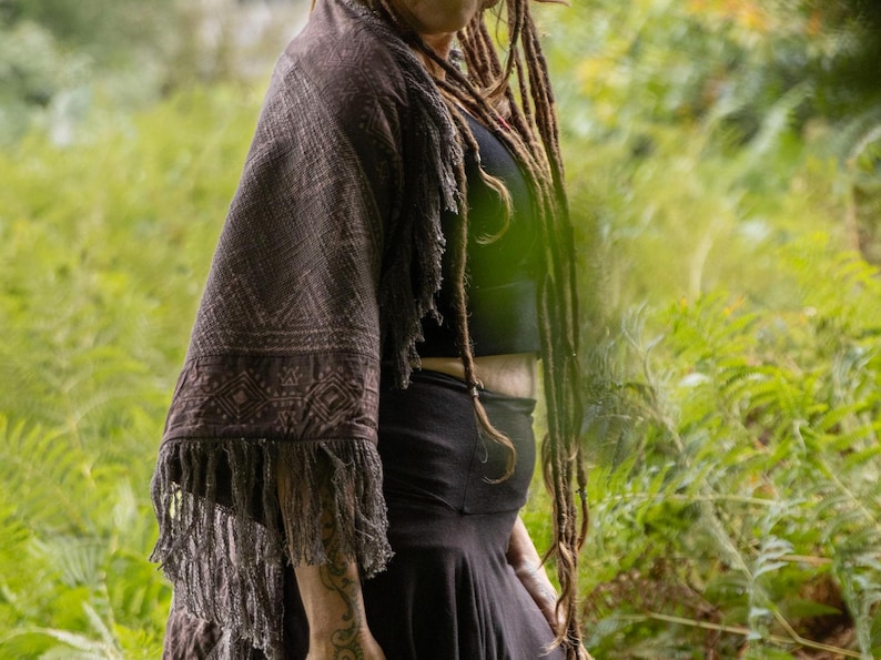 May include: A dark brown shawl with a geometric pattern and fringe is worn over a black top and skirt. The image is set in a green, leafy environment. The shawl's pattern is visible.