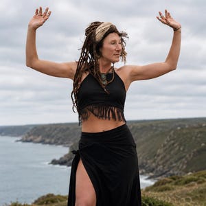 May include: A black halter-style crop top featuring a fringe hemline paired with a matching black wrap skirt that includes a high side slit. The top has a high neckline and the skirt is secured at the waist with a side tie closure.