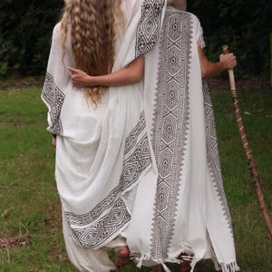 May include: Two people wearing white, flowing garments with geometric patterned trim, standing in a grassy area. One person has long, wavy blonde hair. The other person is wearing a head covering and holding a wooden staff.