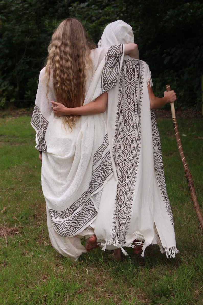 May include: Two people wearing white, flowing garments with geometric patterned trim, standing in a grassy area. One person has long, wavy blonde hair. The other person is wearing a head covering and holding a wooden staff.