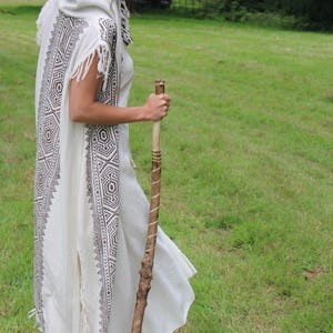 May include: A white hooded robe with geometric patterns in brown and black, worn outdoors. The robe has fringe details and is paired with a wooden walking stick. The person is standing in a grassy field.