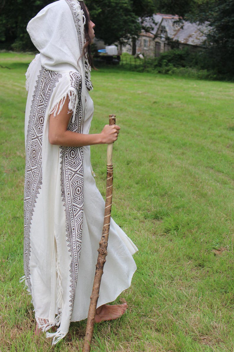May include: A white hooded robe with geometric patterns in brown and black, worn outdoors. The robe has fringe details and is paired with a wooden walking stick. The person is standing in a grassy field.