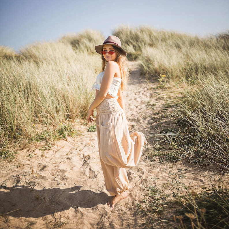 May include: A woman wearing a white crop top, a brown hat, and pink heart-shaped sunglasses stands on a sandy path in a grassy field. She is wearing loose-fitting, light brown pants.
