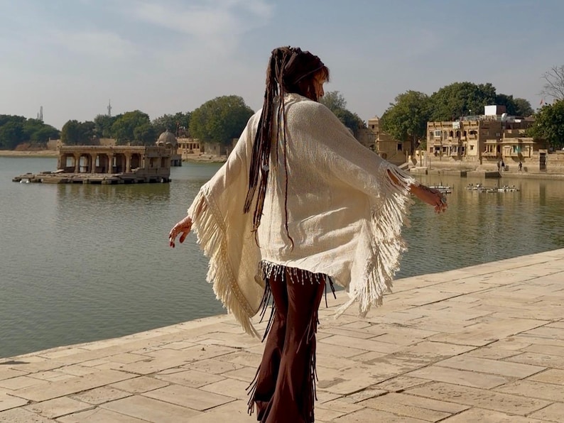 K&ouml;nnte beinhalten: Ein cremefarbener Poncho mit Fransen und einem Perlenbesatz. Der Poncho wird mit braunen Hosen getragen. Der Hintergrund zeigt einen See, einen Steinweg und Geb&auml;ude unter sonnigem Himmel.