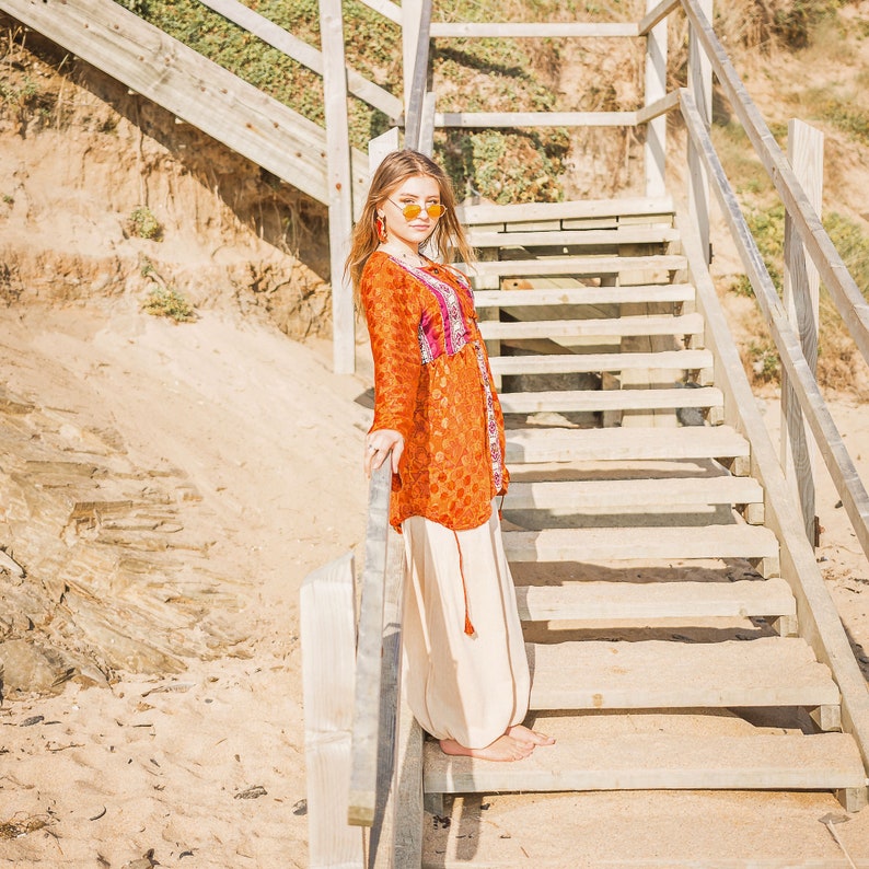 May include: A woman wearing a long, orange and pink patterned tunic with a white, flowing skirt. She is standing on a wooden staircase with a railing. She is wearing sunglasses and has long, dark hair.