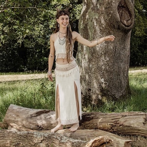 May include: A woman wearing a white crochet top and a long white skirt with a side slit. She is standing on a fallen log in a forest setting.