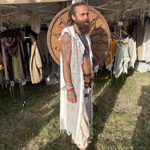 May include: A cream-colored, sleeveless, fringed vest with a geometric pattern. The vest is worn over a matching cream-colored outfit. The background includes a wooden circular design and clothing displayed on racks.