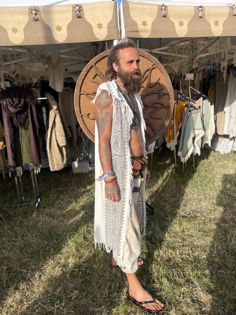 May include: A cream-colored, sleeveless, fringed vest with a geometric pattern. The vest is worn over a matching cream-colored outfit. The background includes a wooden circular design and clothing displayed on racks.