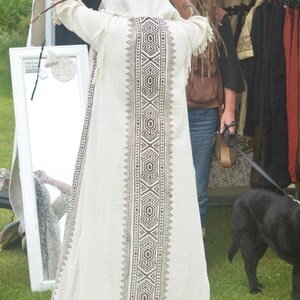 May include: A cream-colored hooded robe with a geometric pattern down the center and fringe at the hem. The robe is being held up by a person. Other clothing items are visible on racks in the background.