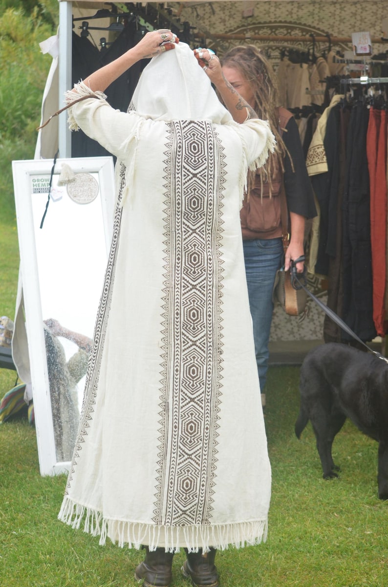 May include: A cream-colored hooded robe with a geometric pattern down the center and fringe at the hem. The robe is being held up by a person. Other clothing items are visible on racks in the background.