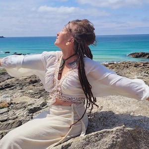 May include: A person wearing a white blouse with embroidered details and matching trousers stands on a rocky beach with arms outstretched. The ocean and blue sky are in the background. They have dreadlocks and are wearing jewellery.