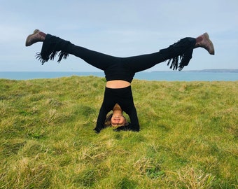 May include: A woman in black pants and a black top does a handstand in a grassy field. She is smiling and has her hair down.