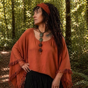 May include: A rust-coloured, flowing top with fringed sleeves and a matching headband. The outfit includes a black skirt and a long, beaded necklace. The woman is in a forest setting.