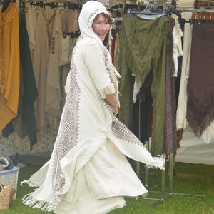 May include: A person wearing a cream-colored, long, flowing robe with intricate brown geometric patterns. The robe has a hood and fringe details. The person is wearing dark brown boots and is standing in front of a clothing rack with various garments.