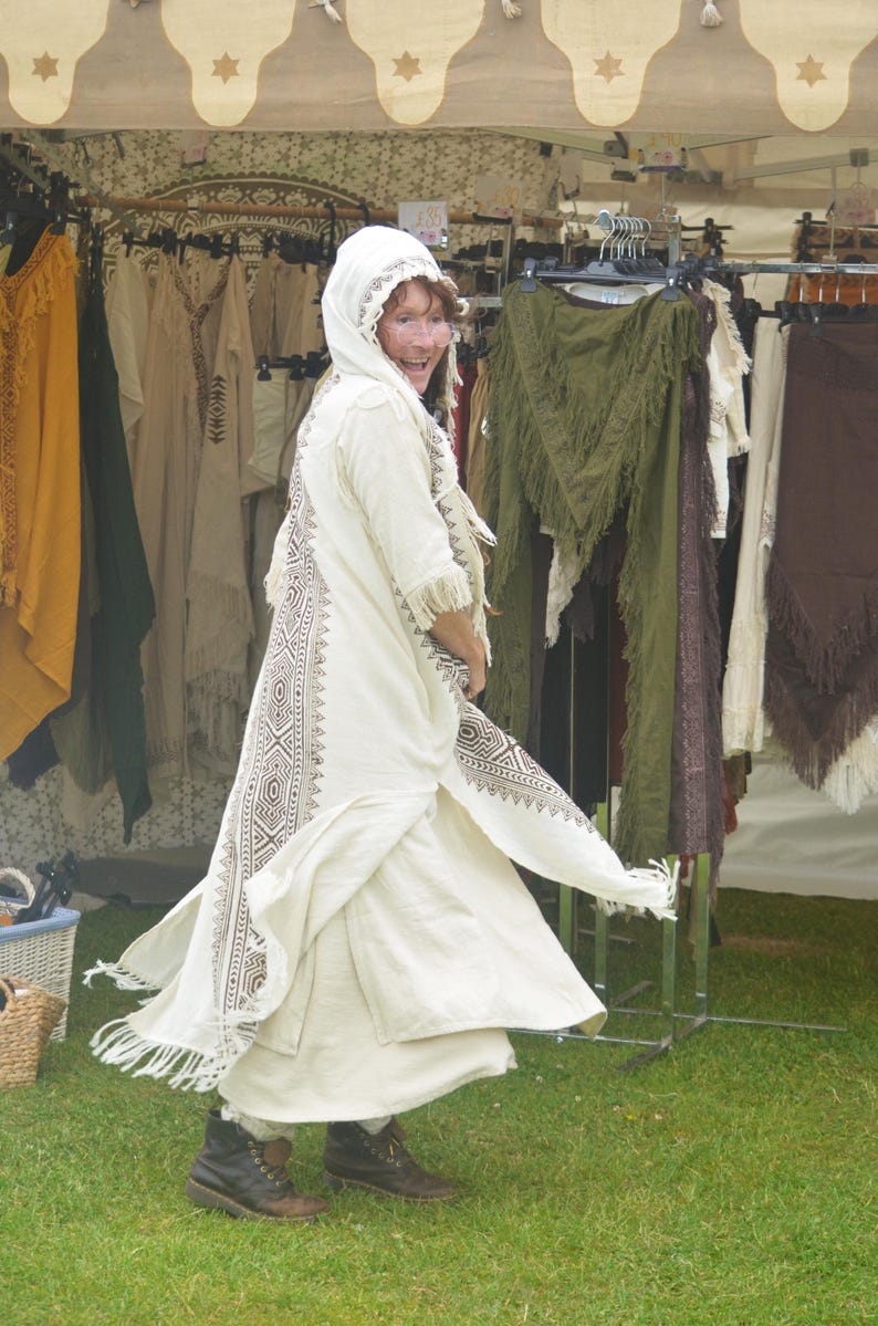 May include: A person wearing a cream-colored, long, flowing robe with intricate brown geometric patterns. The robe has a hood and fringe details. The person is wearing dark brown boots and is standing in front of a clothing rack with various garments.