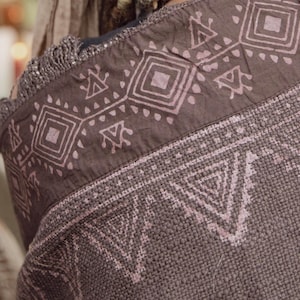 May include: Close-up of a brown tote bag with a geometric pattern in light pink. The bag has a woven texture and a fringed edge. The design features repeating shapes, including squares and triangles, creating a tribal aesthetic.