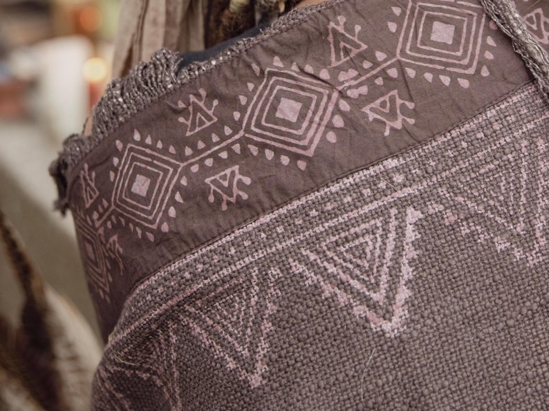 May include: Close-up of a brown tote bag with a geometric pattern in light pink. The bag has a woven texture and a fringed edge. The design features repeating shapes, including squares and triangles, creating a tribal aesthetic.