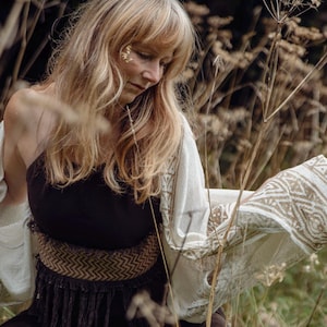 May include: A woman with long blonde hair wears a white crocheted shawl with brown geometric patterns. She is wearing a black sleeveless top with a brown woven belt and fringe. The woman is standing in a field of tall, dry grasses.