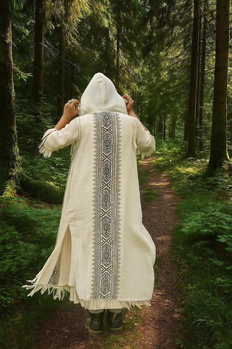 May include: A cream-colored hooded robe with a detailed black and white geometric design down the back. The robe has fringe at the sleeves and hem. The person is walking on a path through a lush green forest.
