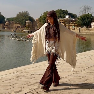 K&ouml;nnte beinhalten: Eine Frau in einem cremefarbenen Schal mit Fransen, einem braunen Oberteil und einer braunen Schlaghose. Sie steht auf einem Steinweg mit einem See und Geb&auml;uden im Hintergrund. Das Outfit hat einen Boho-Stil.