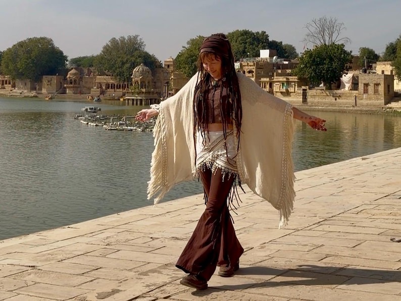 K&ouml;nnte beinhalten: Eine Frau in einem cremefarbenen Schal mit Fransen, einem braunen Oberteil und einer braunen Schlaghose. Sie steht auf einem Steinweg mit einem See und Geb&auml;uden im Hintergrund. Das Outfit hat einen Boho-Stil.