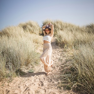 May include: A woman wearing a white crop top, beige pants, a brown hat, and pink sunglasses is standing on a path in a grassy field.