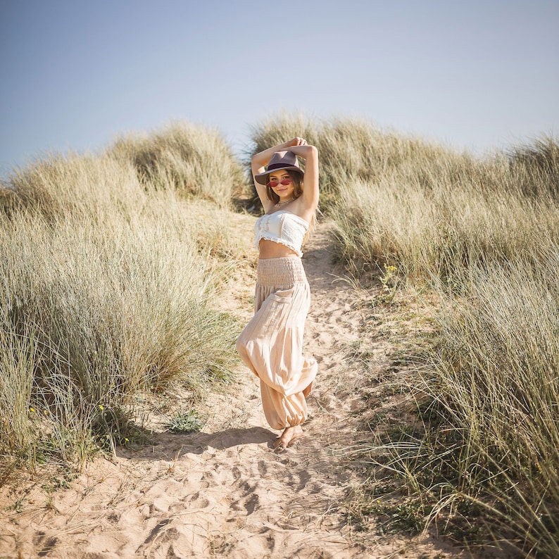 May include: A woman wearing a white crop top, beige pants, a brown hat, and pink sunglasses is standing on a path in a grassy field.