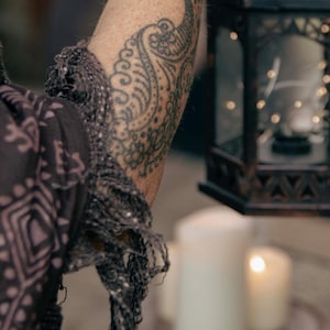 May include: A decorative black metal lantern with a geometric design, holding string lights. A patterned scarf and a tattooed arm are in the foreground. Lit candles are in the background, creating a warm ambiance.