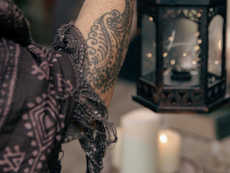 May include: A decorative black metal lantern with a geometric design, holding string lights. A patterned scarf and a tattooed arm are in the foreground. Lit candles are in the background, creating a warm ambiance.